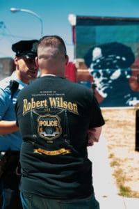 Man wearing a commemorative tshirt for Sgt Robert Wilson, speaking to a police officer with the mural in the background