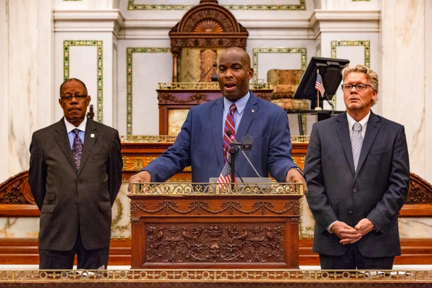 Councilmember Derek Green speaking at the podium during a council session with Commerce Director Harold T. Epps and the Chamber of Commerce for Greater Philadelphia CEO Rob Wonderling looking on.