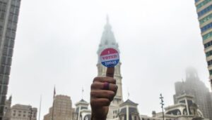 Hand holding an "I voted sticker" in the foreground with Philadelphia City Hall in the background