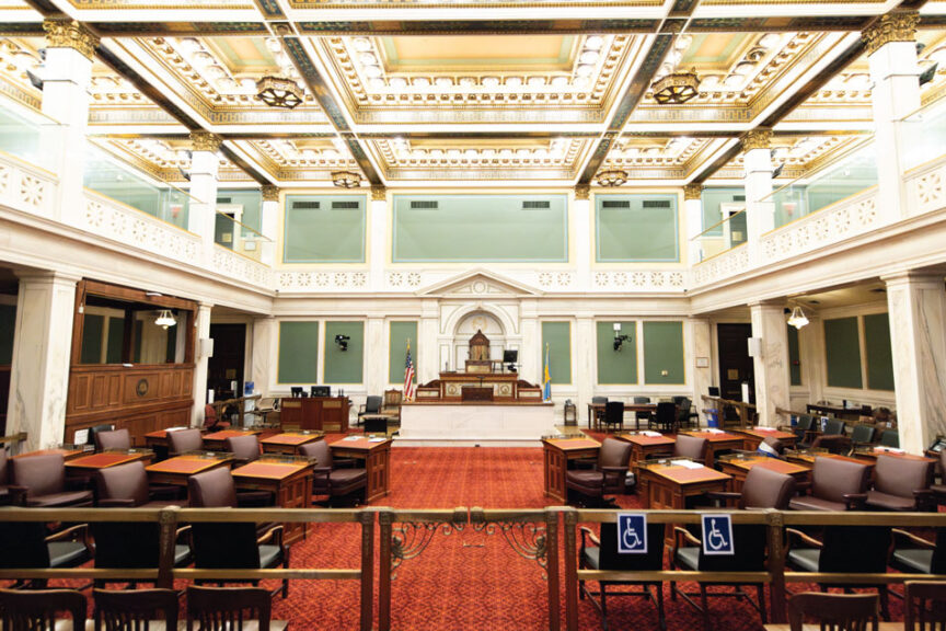 An empty, ornate Philadelphia City Council chamber viewed from behind the spectator gallery. The room features a red carpet with a circular pattern, dark wood desks for councilmembers, and an elevated white marble dais at the front. The American and Philadelphia flags stand behind the main desk. The high ceiling is decorated with intricate gold-trimmed recessed panels and classical architectural molding. Blue wheelchair-accessible signs are visible on the backs of the wooden chairs in the foreground.