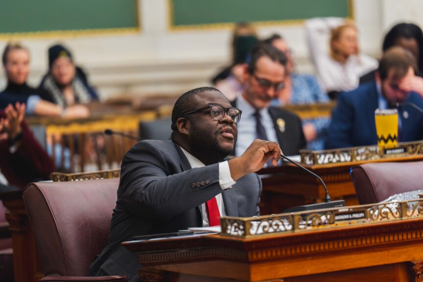Councilmember Dr. Anthony Phillips, wearing a dark suit and glasses, speaks into a microphone while seated at his ornate wooden desk in the Philadelphia City Council chamber. Other council members and staff are visible in the blurred background of the historic room.