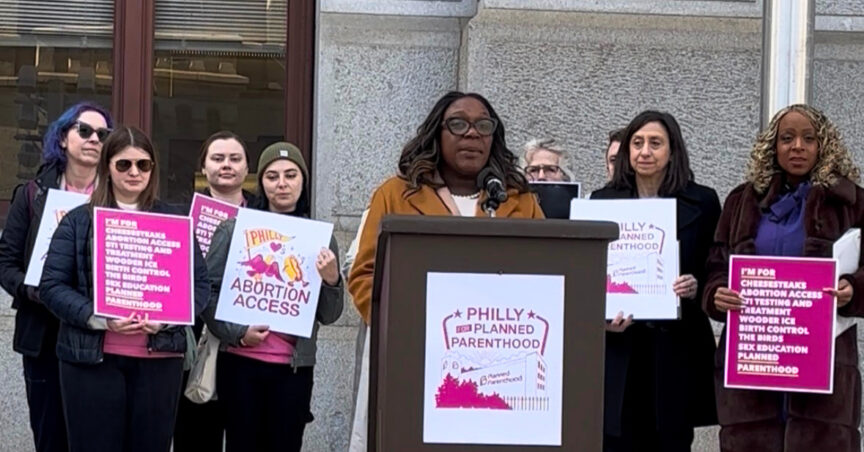 Councilmember Kendra Brooks stands at a wooden lectern with the Philadelphia City seal, speaking during an outdoor press conference. She is wearing a dark patterned blazer and glasses. To her left, two women stand behind her holding signs; one sign reads "Abortion Access Now" and the other features the Planned Parenthood logo with the text "Philly Loves Planned Parenthood." The event is held in front of a historic stone building.