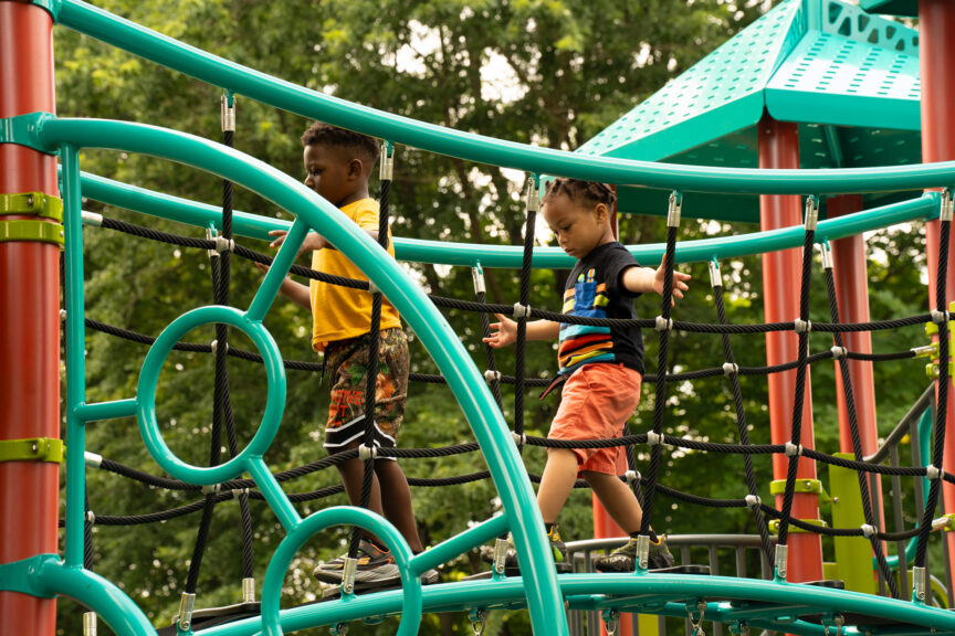 Two young boys climbing on a modern green and black rope course at an outdoor playground, with lush green trees in the background.