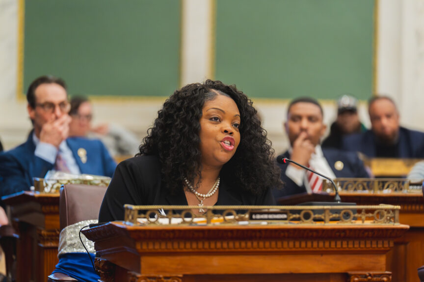 Majority Leader Katherine Gilmore Richardson, wearing a black blazer and a gold "K" necklace, speaking into a microphone at her wooden dais in the ornate Philadelphia City Council chamber.